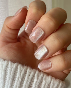 A close-up of a hand displaying short squoval nude shimmer press on nails, with a white knit sweater visible at the cuffs in a cozy lifestyle setting.