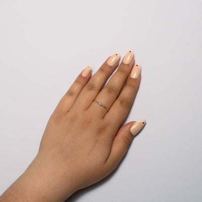 A person's hand resting on a plain white surface, showcasing medium oval nude press on nails with a white french tip and a tiny red heart accent at the cuticle. A delicate silver ring with a small light-colored round stone is worn on the middle finger.