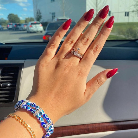 A hand displaying short almond red press on nails wearing a diamond ring and blue evil eye bracelets, photographed inside a car with a parking lot view in the background.