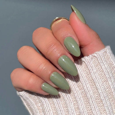 A close-up of a hand displaying short almond green press on nails, wearing a gold ring and a beige knit sweater against a gray-blue background.