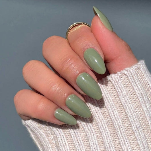 A close-up of a hand displaying short almond green press on nails, wearing a gold ring and a beige knit sweater against a gray-blue background.