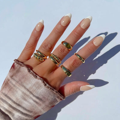 A hand wearing short almond nude press on nails with a classic white french tip, accented with chunky gold rings, rests against a soft pink knit sweater background.