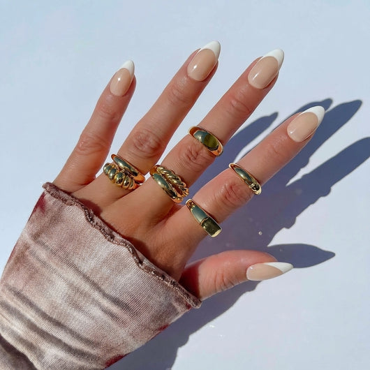 A hand wearing short almond nude press on nails with a classic white french tip, accented with chunky gold rings, rests against a soft pink knit sweater background.