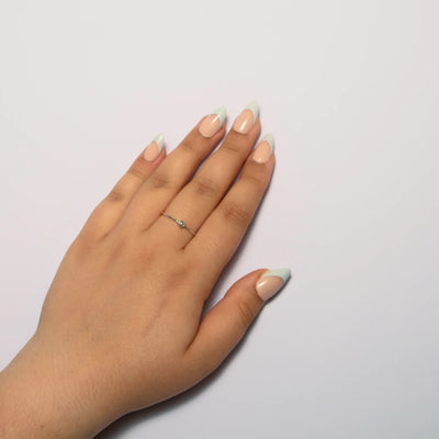 A person's hand resting on a plain white surface, showcasing short almond nude press on nails with a sage green french tip. A delicate silver ring with a small light-colored round stone is worn on the middle finger.