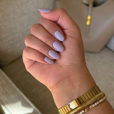 A hand with deep skin tone displaying short oval lavender press on nails in a lifestyle setting, paired with a gold watch and bracelet against a beige cushioned background.