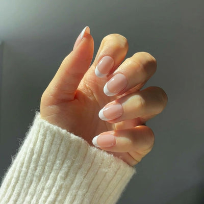A close-up of a hand displaying short oval nude french tip press on nails, with a white knit sweater cuff visible against soft natural lighting.