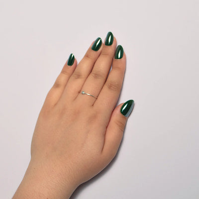 A person's hand resting on a plain white surface, showcasing short oval green press on nails. A delicate silver ring with a small light-colored round stone is worn on the middle finger.