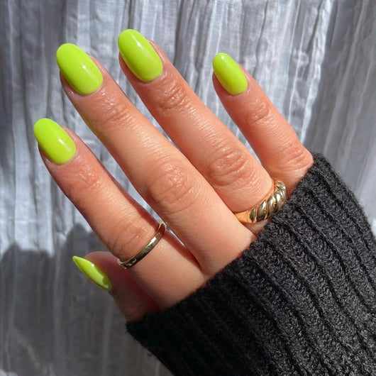 A close-up of a hand displaying short oval green press on nails, with gold rings on the index and middle fingers, holding a black knit fabric in a lifestyle setting.