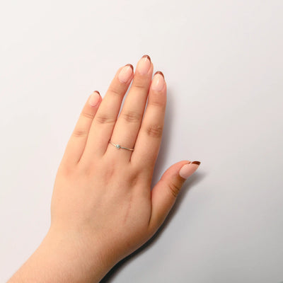 A person's hand resting on a plain white surface, showcasing short oval nude french tip press on nails. A delicate silver ring with a small light-colored round stone is worn on the middle finger.