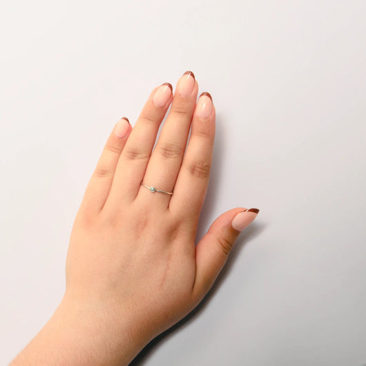A person's hand resting on a plain white surface, showcasing short oval nude french tip press on nails. A delicate silver ring with a small light-colored round stone is worn on the middle finger.