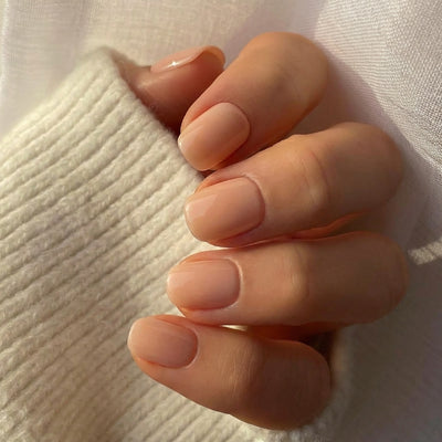 A close-up of a hand displaying short squoval nude press on nails, with a soft white knit sweater visible in a cozy lifestyle setting.