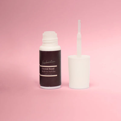 Lashnetix Crystal Bond brush-on nail glue, 7ml / 0.24 fl oz, with the cap removed to reveal the brush applicator, photographed against a soft pink background.